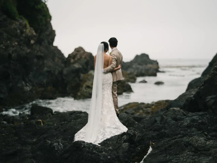 Bride and groom embrace on dark rocks with ocean and forested shoreline beyond at Black Rock Oceanfront Resort