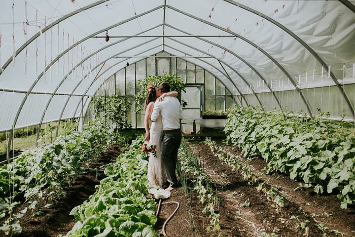 Couple embracing inside a glass greenhouse at Duck Creek Farm, surrounded by green plants and garden beds