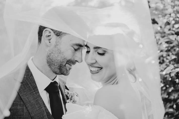 Bride and groom in white fabrics embrace outdoors in bright natural light, eyes closed in an intimate moment