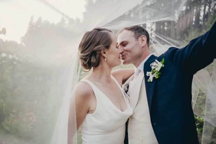 Bride and groom kissing in profile beneath sheer fabric, soft natural light filtering through trees in background