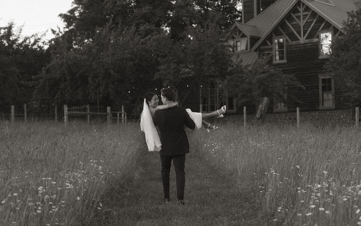 Groom in dark suit carries bride in white veil across grassy field with tree line and house visible behind them at dusk