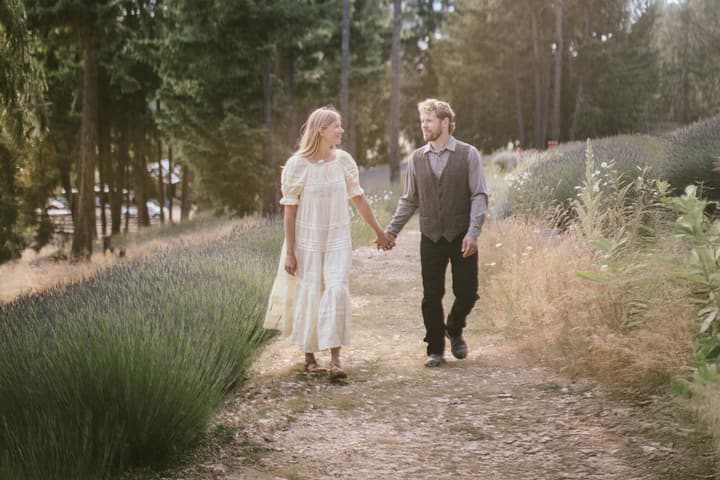 Couple walking hand-in-hand down a gravel path lined with lavender plants and tall evergreen trees in dappled sunlight