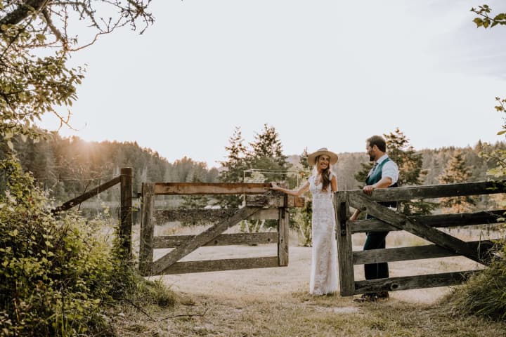 Woman in white lace dress and wide-brimmed hat standing by weathered wooden fence with forested hillside beyond