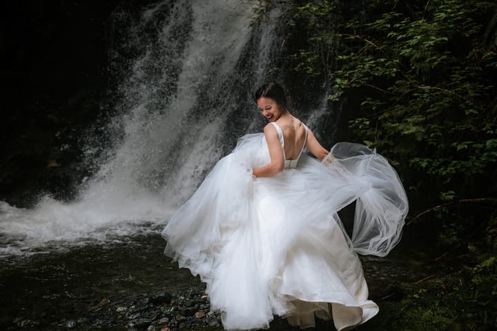 Bride in white tulle gown with open back stands before a weathered concrete wall with cascading water and green foliage