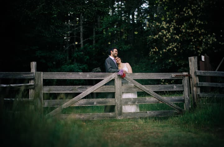 Couple in wedding attire stand at a weathered wooden farm gate with a bouquet, dense forest and water visible behind them