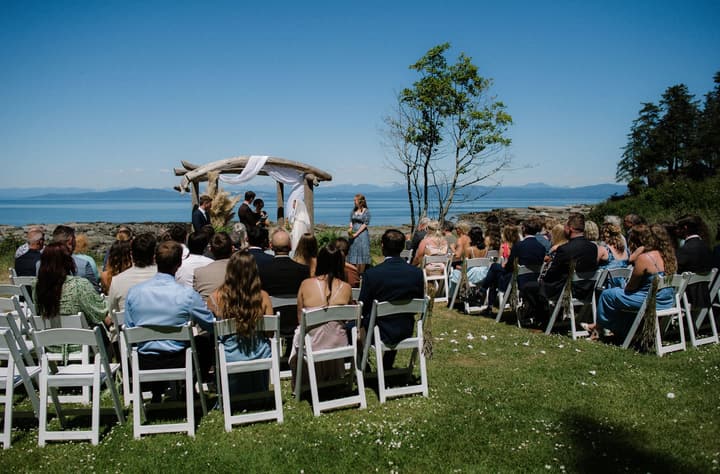Outdoor wedding ceremony on a grass lawn overlooking a blue ocean and distant mountains, with white chairs and a wooden arbor