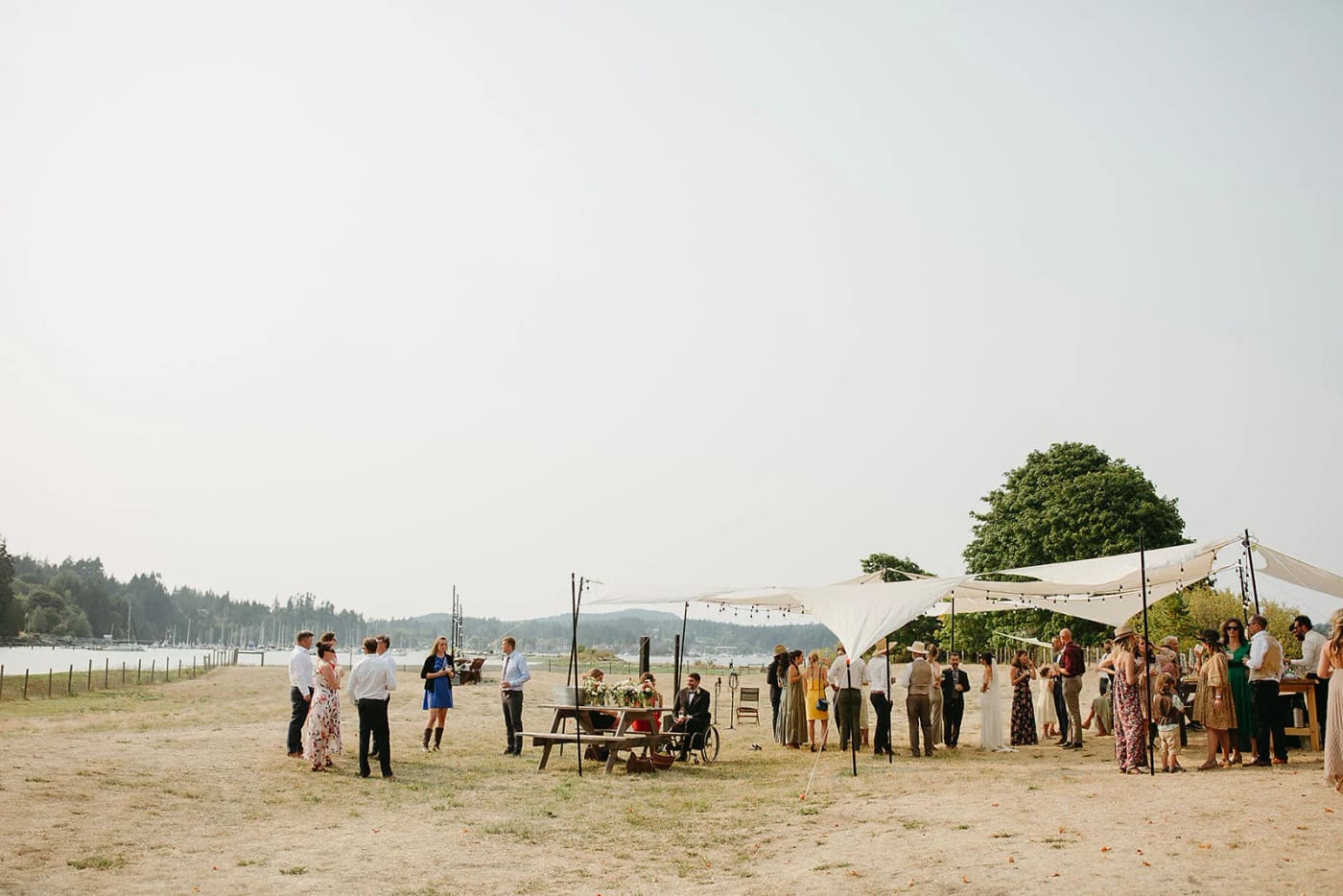Outdoor wedding ceremony under a cream canvas canopy with guests seated on wooden benches, mountains visible across the