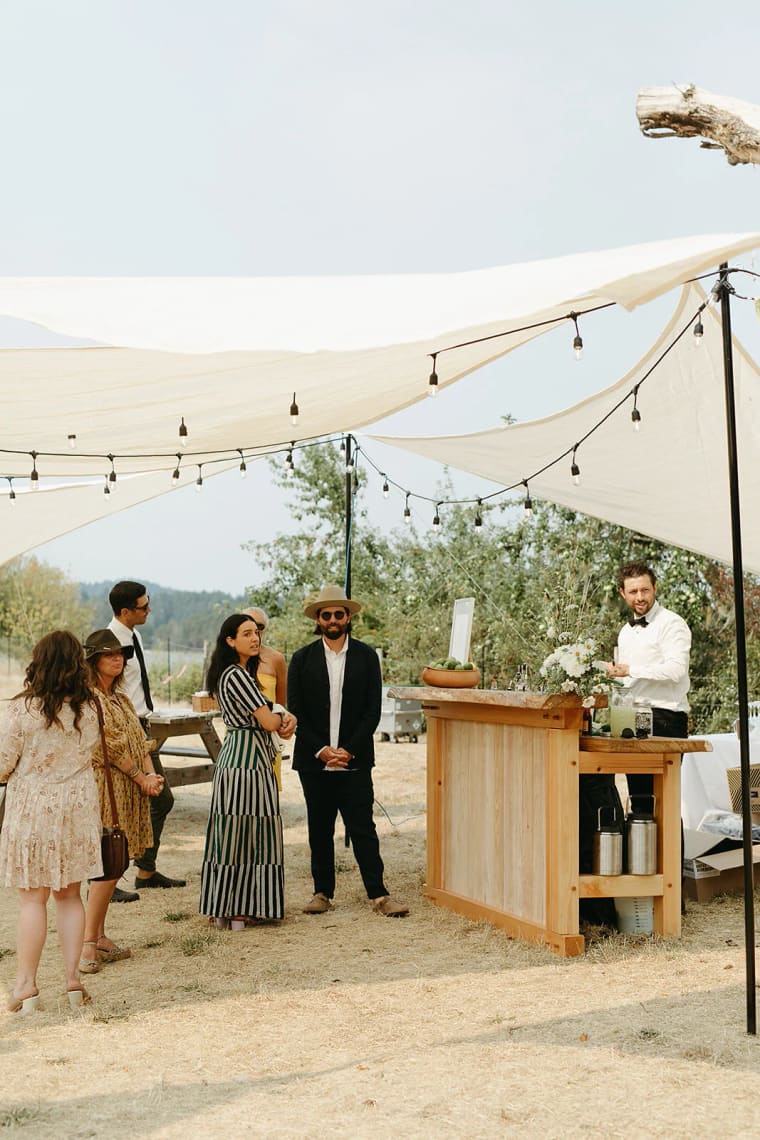 Couple in striped dress and dark blazer stand under cream canvas canopy with hanging string lights