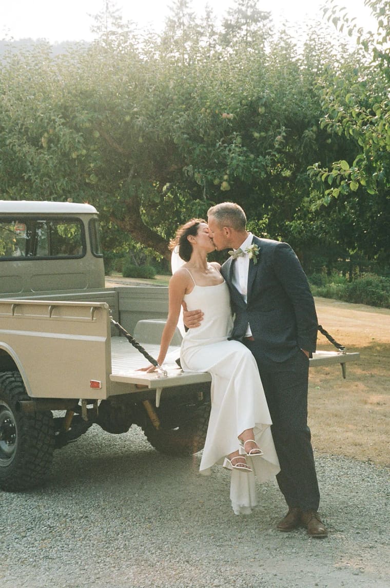 Couple kissing on wooden bench with lush green hedge wall and coniferous trees behind them in afternoon light