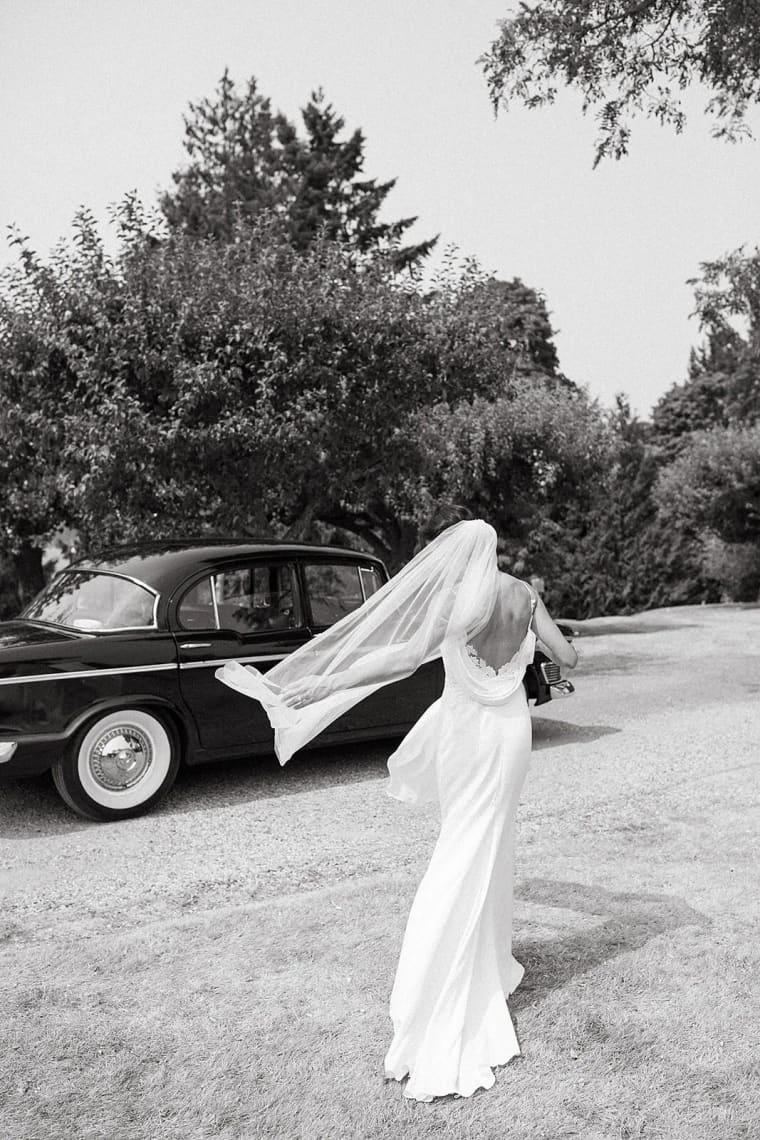Bride in flowing white gown and veil stands on gravel drive beside vintage dark car with trees behind