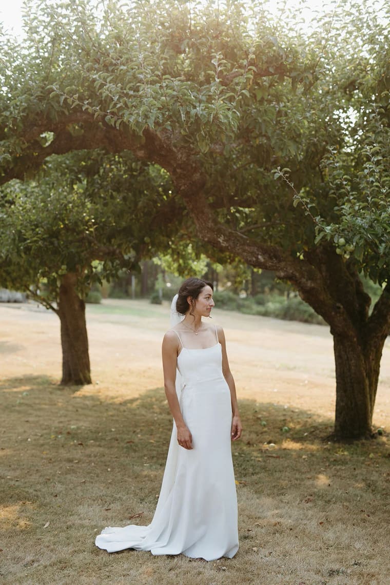 Bride in white sleeveless gown standing on grass beneath large mature tree with trailing branches in afternoon light