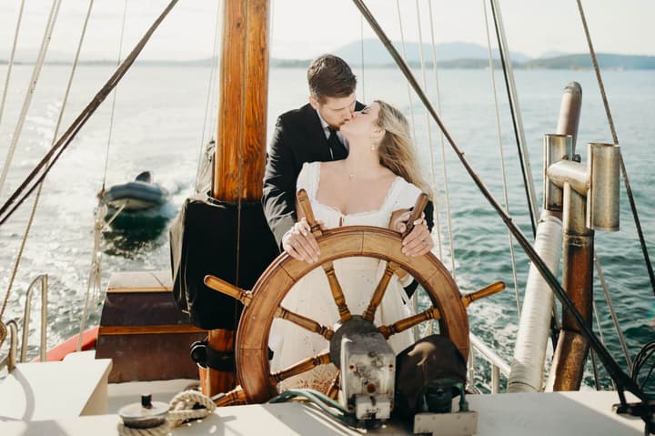 Couple kissing at the helm of a wooden sailing vessel with hands on the ship's wheel