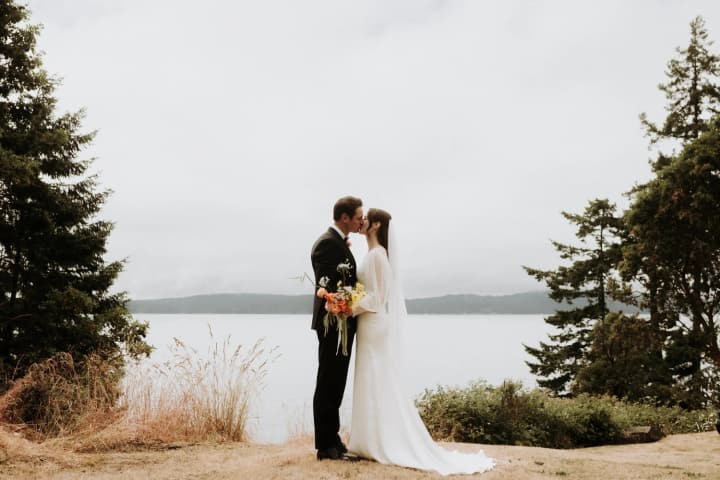 Groom in dark suit and bride in white dress kiss on sandy beach beside calm water, holding wildflower bouquet